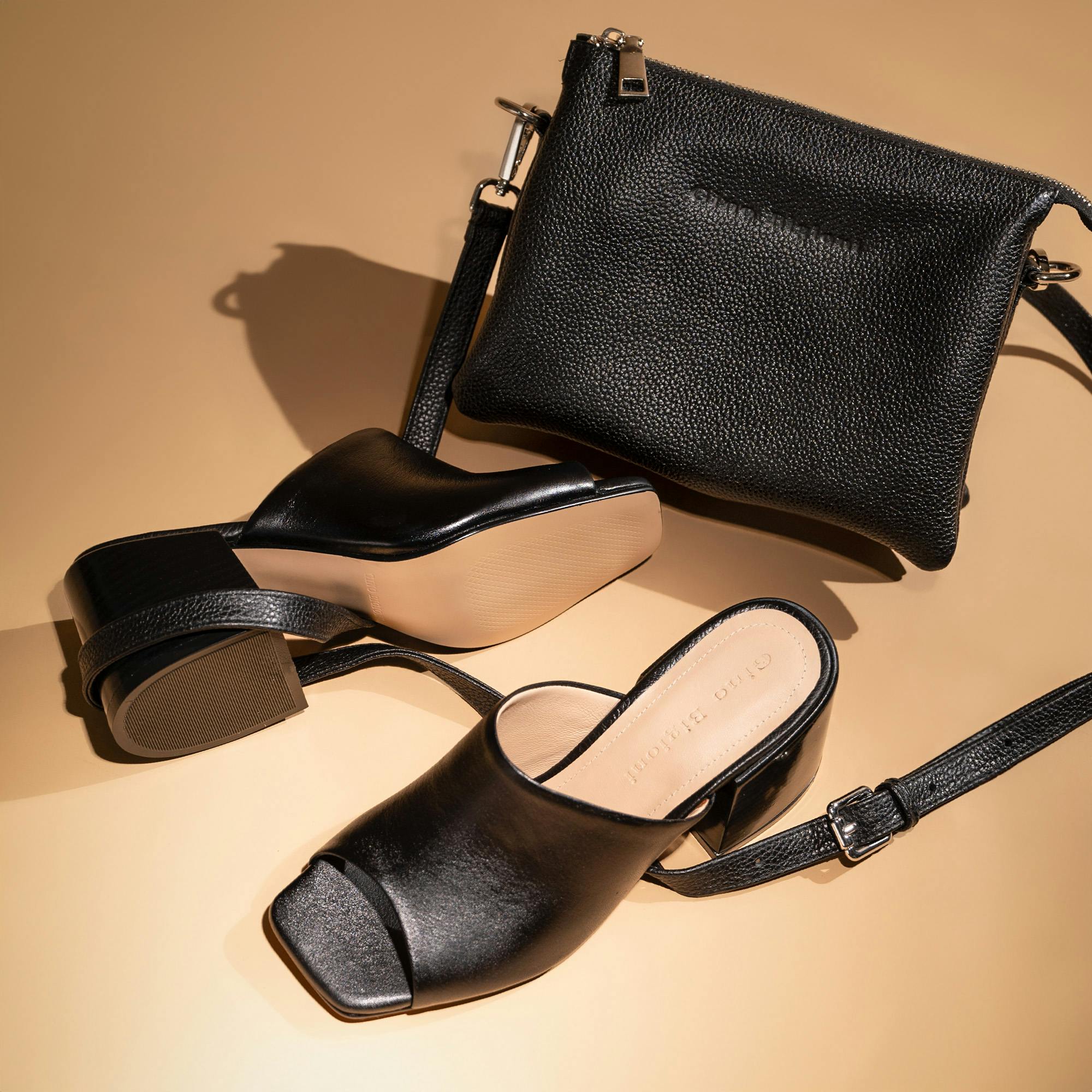 A stylish flat lay featuring a pair of black leather open-toe block heel sandals and a matching textured black leather crossbody bag with a zipper, all arranged on a beige background.